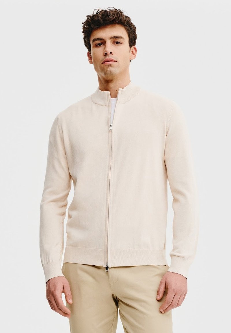 Jasno beżowy sweter na zamek wykonany z miękkiego materiału, z kołnierzem stojącym i prążkowanymi mankietami, w połączeniu z brązowymi spodniami.