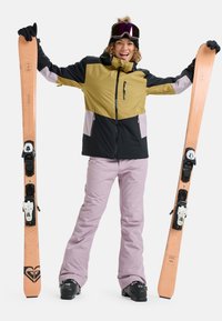 Skiausrüstung mit einer zweifarbigen Jacke (schwarz und oliv) mit Reißverschluss, passenden rosa Hosen und orangefarbenen Skiern mit schwarzen Bindungen und Details.