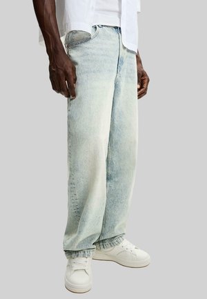 Homme portant un jean décontracté en denim clair associé à des baskets blanches et une chemise blanche partiellement visible sur un fond gris uni.