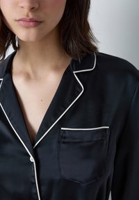 Czarna jedwabna koszula nocna z białym wykończeniem; posiada kieszeń na piersi. Materiał ma gładką fakturę i klasyczny kołnierzyk.