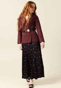 Bordeauxfarbener Blazer mit zwei vorderen Taschen, kombiniert mit einem schwarzen, bodenlangen Rock mit kleinen roten Blumenmustern, getragen mit schwarzen High Heels.
