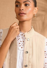 Beige sleeveless vest with scalloped collar, fastened by silver buttons, worn over a white lace blouse with floral patterns. Smooth texture.