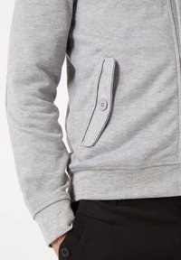 Sudadera gris con cremallera hecha de una tela suave, que presenta dos bolsillos laterales con cierres de botón y puños acanalados. Sin patrones distintivos.