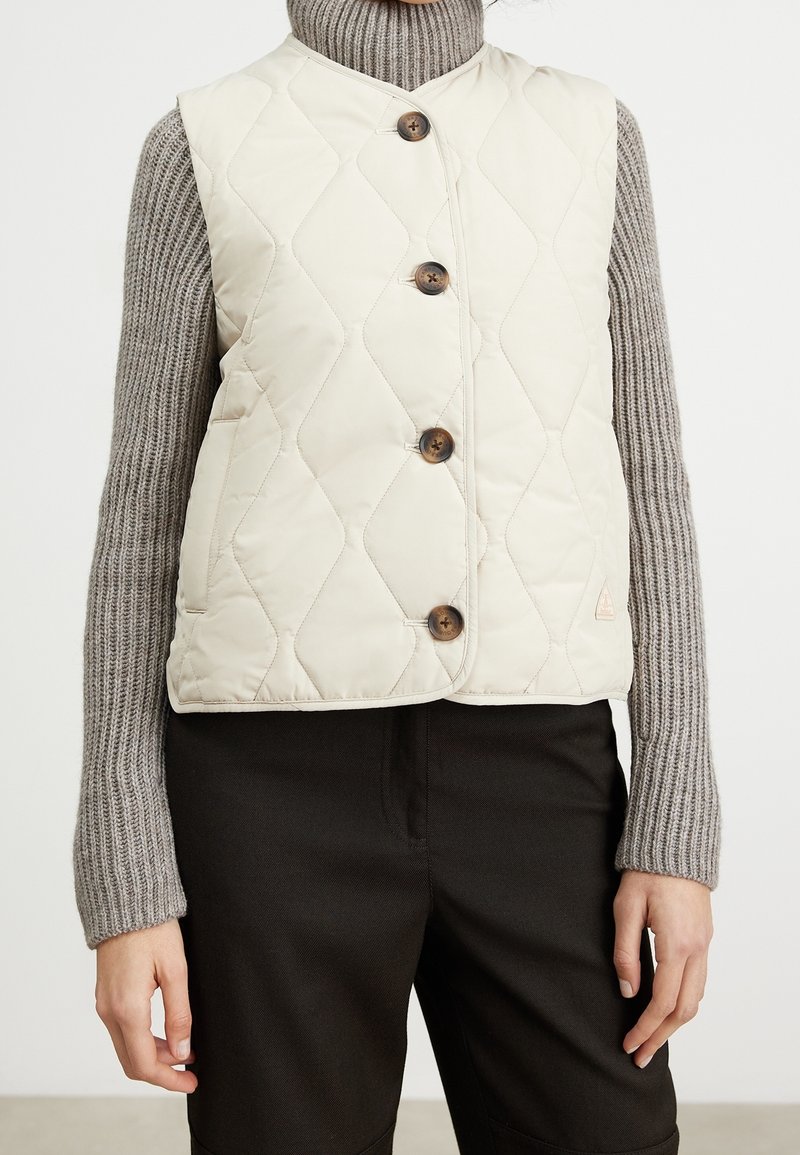 Femme portant une veste matelassée crème avec des boutons en bois par-dessus un pull col roulé gris côtelé et un pantalon noir, debout devant un fond uni.