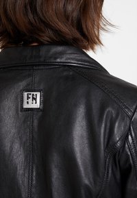 Svart läderjacka med en slät yta, framträdande sömmar och en logotyp med "FN" på baksidan av kragen.