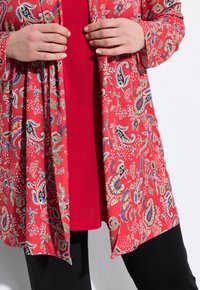 Punainen paisley-kuvioinen auki oleva neuletakki, joka on valmistettu kevyestä kankaasta, kerroksittain yhdenmukaisen punaisen täyspaidan päällä, yhdistettynä mustiin housuihin.