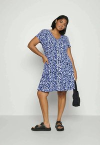 Vestido floral azul com mangas curtas, decote em V e saia em camadas. Modelo usa sandálias pretas e segura uma pequena bolsa preta.