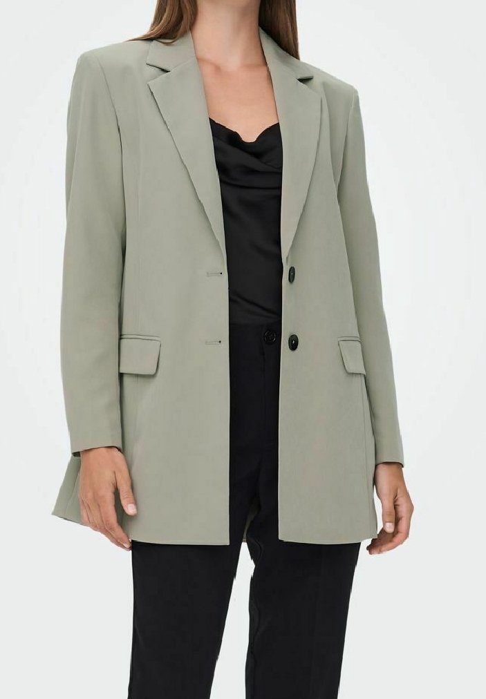Femme portant un blazer gris clair sur mesure, un haut en satin noir et un pantalon noir, debout avec les bras détendus le long du corps.