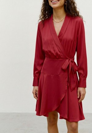 Une femme souriante porte une robe portefeuille en satin bordeaux, à manches longues et nouée à la taille, avec un collier doré.