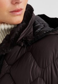 Nahaufnahme einer Person, die eine schwarze gesteppte Jacke mit einem Fleece-Kragen und einen weißen gestrickten Rollkragenpullover trägt.