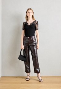 Combinaison en dentelle noire à manches courtes associée à un pantalon en cuir bourgogne brillant taille haute avec des coutures rouges. Le mannequin tient un petit sac à main noir.