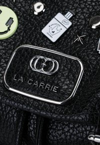 Borsa a tracolla nera con texture, caratterizzata da una placca in metallo argentato con la scritta "LA CARRIE". Distintivi colorati e borchie decorative aggiungono dettagli.