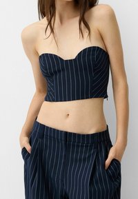 Žena v námořnický modrý pruhovaný topu bez ramínek a odpovídajících kalhotách s vysokým pasem, s rukama v kapsách.