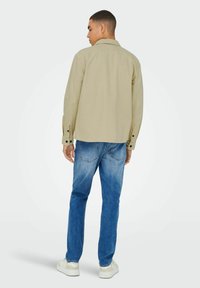 Beige Hemd mit Knöpfen, Kragen, langen Ärmeln und schwarzen Knöpfen, kombiniert mit verwaschenen blauen Jeans und weißen Sneakern.