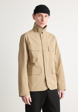 Beige, lightweight jacket with a high collar, snap buttons, and four front pockets. Made of a smooth fabric with a matte finish. Black knit hat.