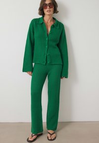 Vihreä neulottu kaksiosainen asu, joka sisältää nappimaisen takin, jossa on kaulus, sekä siihen sopivat slim fit -housut. Kevyt kangas, jossa on tekstuuri.