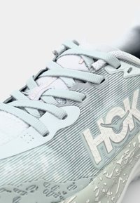 Light blue athletic shoe with textured upper, flat laces, and a prominent white logo. Features a layered sole with a patterned design.