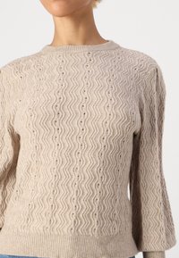 Beige knit sweater with zigzag pattern, round neckline, and ribbed cuffs. Soft texture, semi-fitted shape, and detailed stitching.