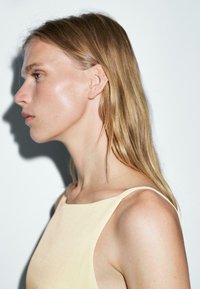 Giovane donna con lunghi capelli biondi che indossa un top senza maniche beige chiaro, rivolta a sinistra su uno sfondo bianco semplice.