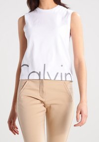 Vit ärmlös topp med rund halsringning och grå "Calvin"-logotyp tryckt på framsidan. I kombination med beige figurnära byxor med sidofickor.