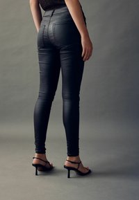 Jeansi skinny negri cu talie înaltă, cu o textură fină, având buzunare la spate și cusături subtile, asortați cu sandale negre cu toc înalt și curele.