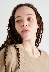 Suéter de punto de color marrón claro con cuello redondo, que presenta una tela texturizada. El modelo tiene el cabello rizado y largo, y un piercing en el septum de plata.