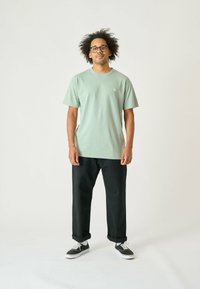 Hellgrünes Baumwoll-T-Shirt mit kurzen Ärmeln, Rundhalsausschnitt, kleinem Logo auf der Brust, kombiniert mit lockeren schwarzen Hosen und Sneakern.