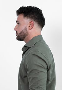 Olijfgroene shirt met een button-down kraag, lange mouwen en een gladde textuur; zijkant getoond om de pasvorm en het ontwerp te accentueren.