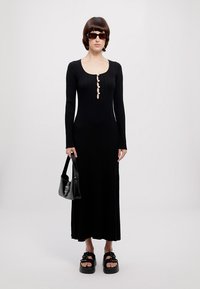 Black ribbed long dress with buttoned front, long sleeves, paired with a black handbag and chunky platform sandals. Flat background.