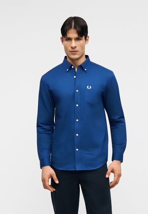 Homme portant une chemise bleu ajustée à manches longues avec une poche poitrine et un pantalon sombre, debout devant un fond uni.