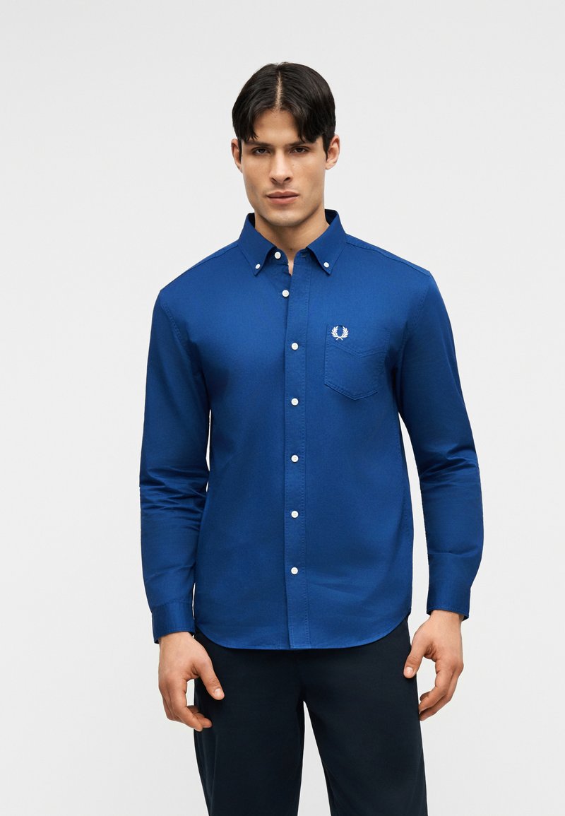 Homme portant une chemise bleu ajustée à manches longues avec une poche poitrine et un pantalon sombre, debout devant un fond uni.