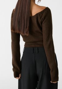 Femme aux longs cheveux bruns lisses portant un pull en tricot marron à épaules dénudées et un pantalon noir taille haute, vue de dos.
