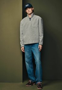 Mladý muž oblečený v šedom svetri s krátkym zipsom, modrých džínsach, tmavohnedých topánkach a námořníckej čiapke stojí pred dvojfarebnou zelenou stenou.