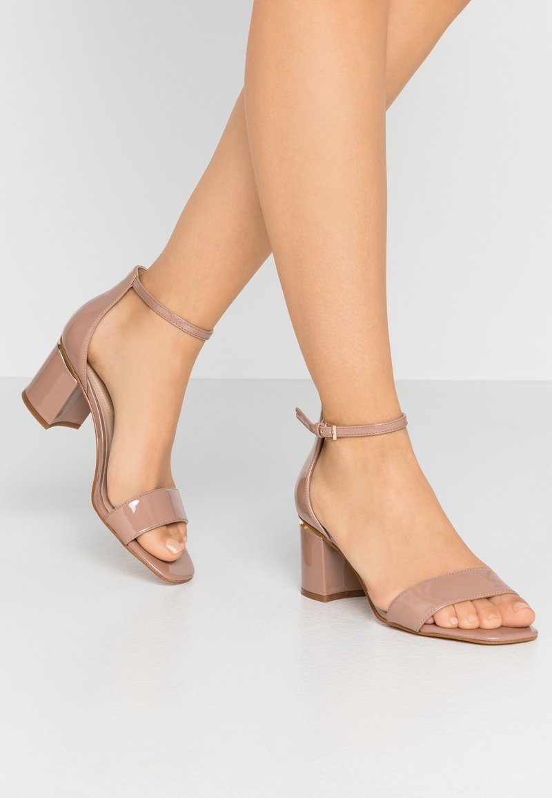 Jambes portant des sandales à talon bloc en cuir verni couleur nude avec des brides à la cheville sur un sol et un fond gris clair.