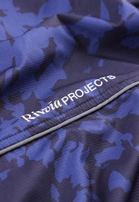 Ύφασμα σε αποχρώσεις του μπλε και σκούρου μωβ με λευκή επιγραφή "Rivūōia PROJECTS" και αντανακλαστική γκρι ραφή λεπτομέρειας.