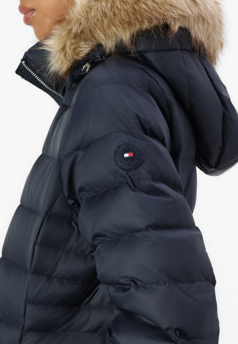 Marineblaue Puffy-Jacke mit einem mit Fell gefütterten Kapuze, gestepptem Material und einem runden Logo-Patch auf dem linken Ärmel. Mit Reißverschluss vorne zu schließen.