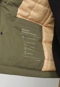 Green outerwear jacket interior with tan quilted lining, featuring a label that lists material and performance details. Textured fabric.