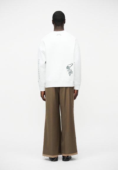 Άνδρας που φοράει λευκό φούτερ με μαύρο σχέδιο χεριού και λουλουδιού στην πλάτη, και καφέ παντελόνι με φαρδιά μπατζάκια και μπεζ μανσέτες, στέκεται γυρισμένος προς τα πίσω.