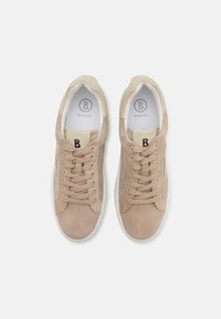 Beige semsket sko med en glatt skinnstunge, snøring og hvite gummisåler. Har en svart logo på stungen.