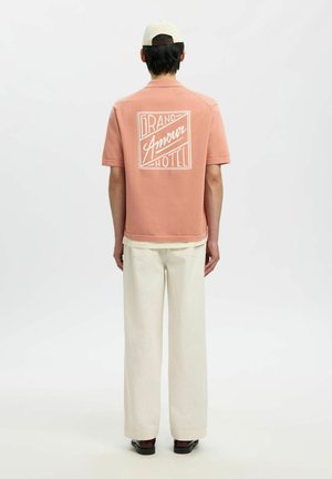 Osoba v ružovej krátkemu rukávu s nápisom „Grand Amour Hotel“ na chrbte, bielymi širokými nohavicami, bielou šiltovkou a čiernymi topánkami, stojaca.