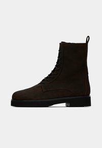 Botte chelsea en cuir marron avec un bout arrondi, un laçage à l'avant et un talon renforcé. Elle présente une semelle noire épaisse et une languette à l'arrière.