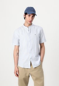 Camicia a maniche corte con bottoni a righe blu chiaro e bianche, con colletto rialzato, abbinata a un cappellino blu. I pantaloni beige completano l'outfit.