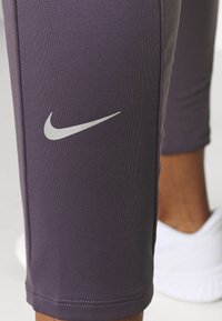 Nike legíny do půli lýtek v tmavě fialové barvě, s logem stříbrného swoosh, těsný střih, hladká textura a lemování těsně pod kolenem.