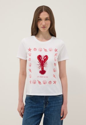 Donna che indossa una maglietta bianca con un design di aragosta rossa e conchiglie, con la scritta "Vita sotto il mare", abbinata a jeans blu.