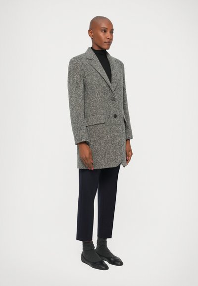 Houndstooth mintázatú szürke kabát egy gombbal, oldalsó zsebekkel és kifordított hajtókával, sötétkék nadrággal és fekete lapos cipőkkel párosítva.