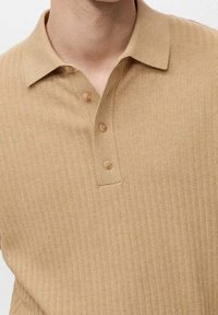 Polo beige à côtes fabriqué en tissu texturé, doté d'un col et de trois boutons de couleur assortie le long de la patte de boutonnage.