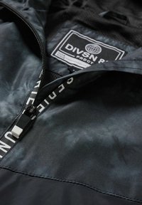 Chaqueta negra y gris con una textura suave y brillante. Presenta un cierre frontal, forro de malla y una etiqueta con el texto "DIVSN 81 PRODCT."