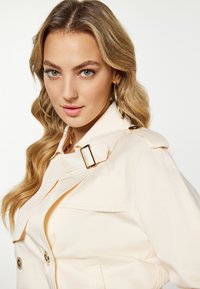 Lyscreme trenchcoat med guld hardware detaljer. Har en bæltestrop ved kraven, store knapper og en glat, elegant tekstur.
