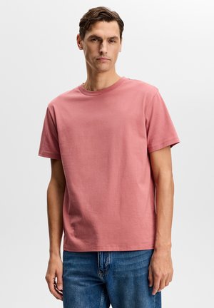 Homme portant un t-shirt à manches courtes uni rose poussiéreux et un jean bleu, debout devant un fond clair neutre.