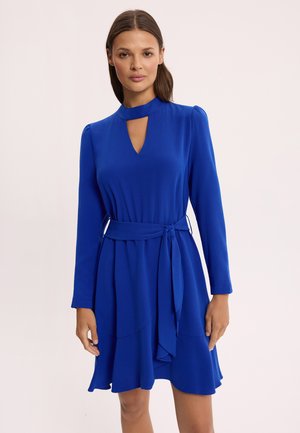 Kobieta w długiej sukience z długim rękawem w królewskim błękicie, z dekoltem w kształcie łezki oraz paskiem wiązanym w talii, stojąca na jednolitym jasnym tle.